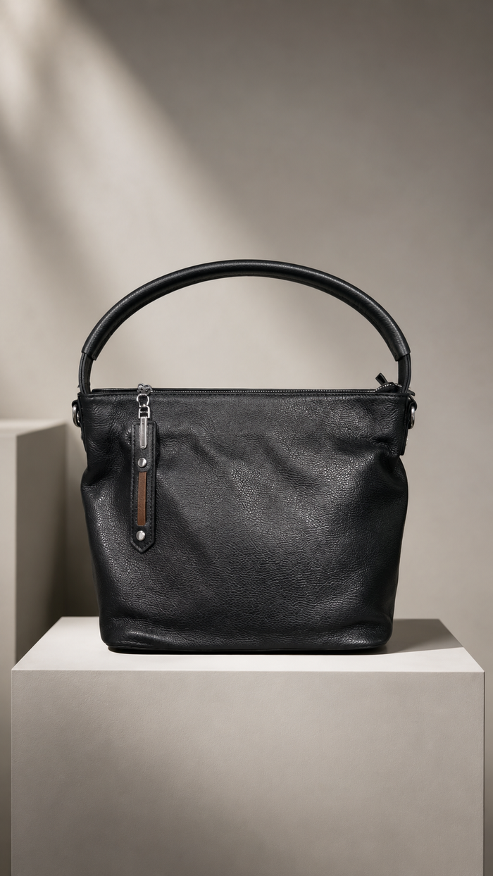 A black textured leather handbag with a curved top handle, displayed on a minimalist beige pedestal against a neutral background.