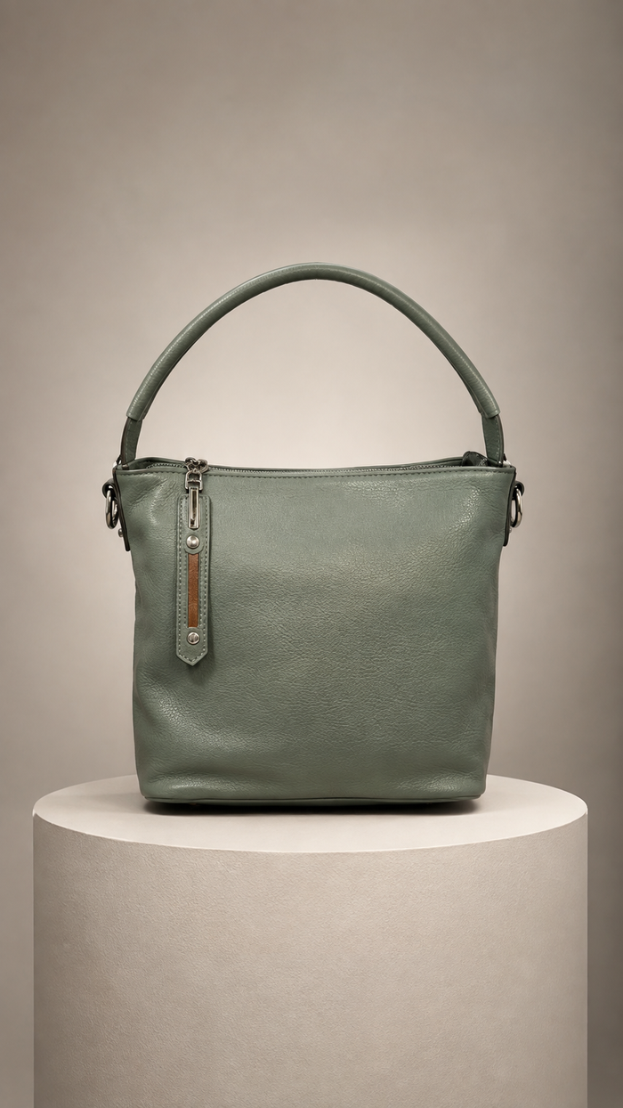 A sage green leather bucket bag with a top handle, displayed on a round pedestal against a neutral beige background.