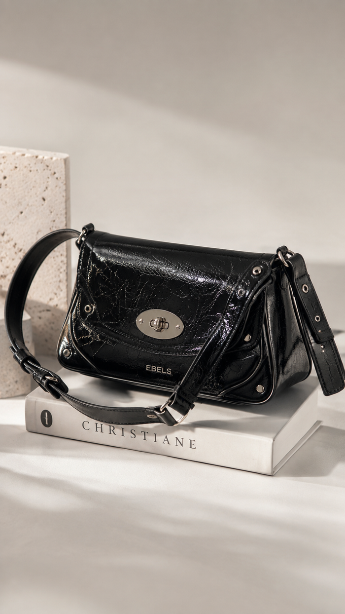 A black glossy crocodile-embossed leather shoulder bag by Ebels, styled on top of a white book with 'CHRISTIANE' printed on it, set against a neutral beige background.